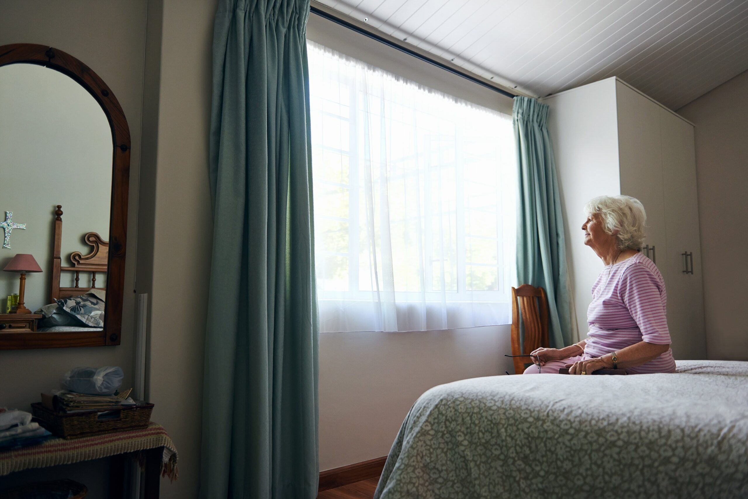 Elderly Woman Sitting On Bed Looking Out Window